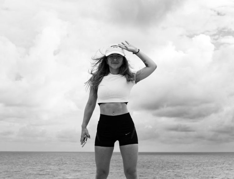 Woman in sportswear stands confidently by the sea against a cloudy sky.