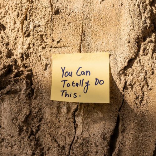 A sticky note with a motivational message on a textured wall in Dubai, with sunlight casting shadows.
